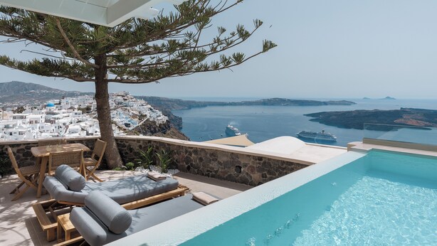 A poolside area with lounge chairs, a table, and a view of a coastal town and the sea. - Morfes Luxury Suites by Caldera Houses (Santorini)