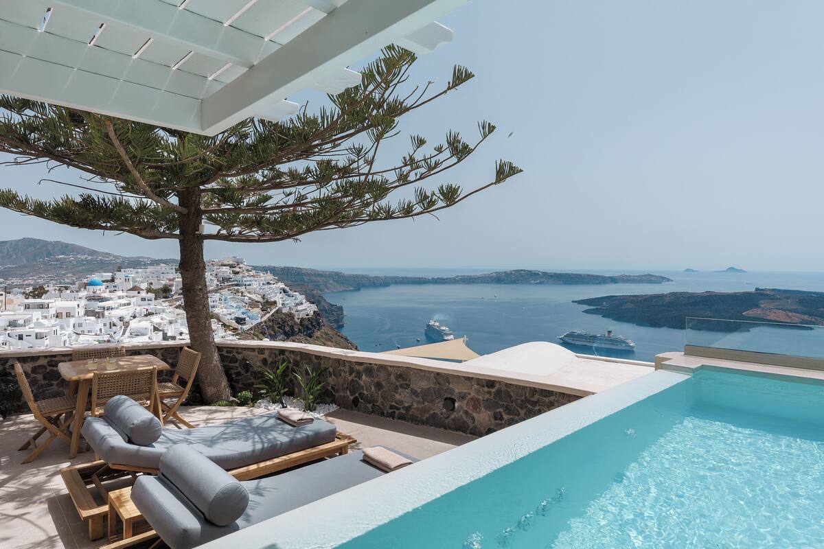 Zona de piscina con sillones, una mesa y vistas a un pueblo costero y al mar.
