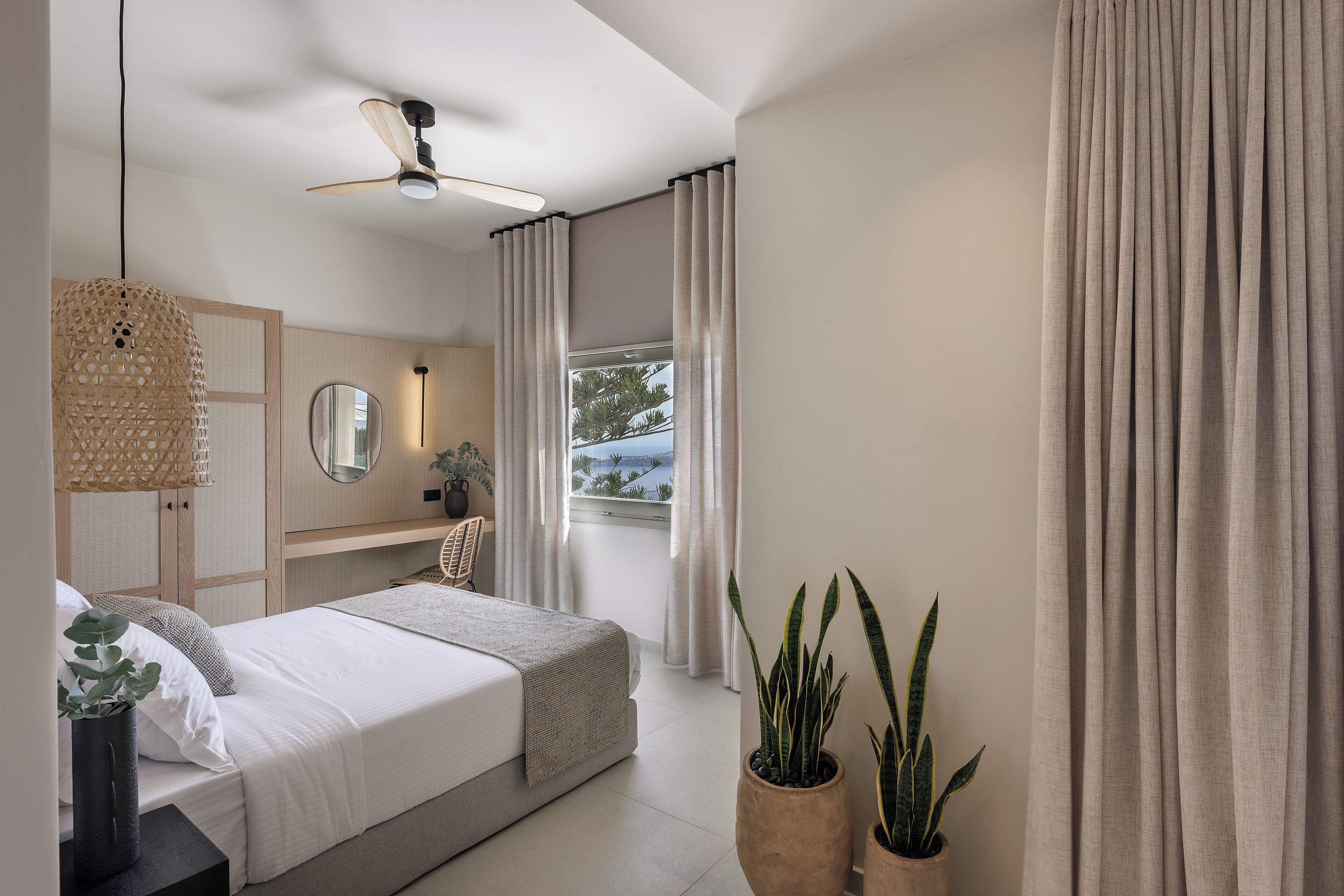 Un dormitorio moderno con cama, mesita de noche, espejo, ventilador de techo y una ventana grande con cortinas.