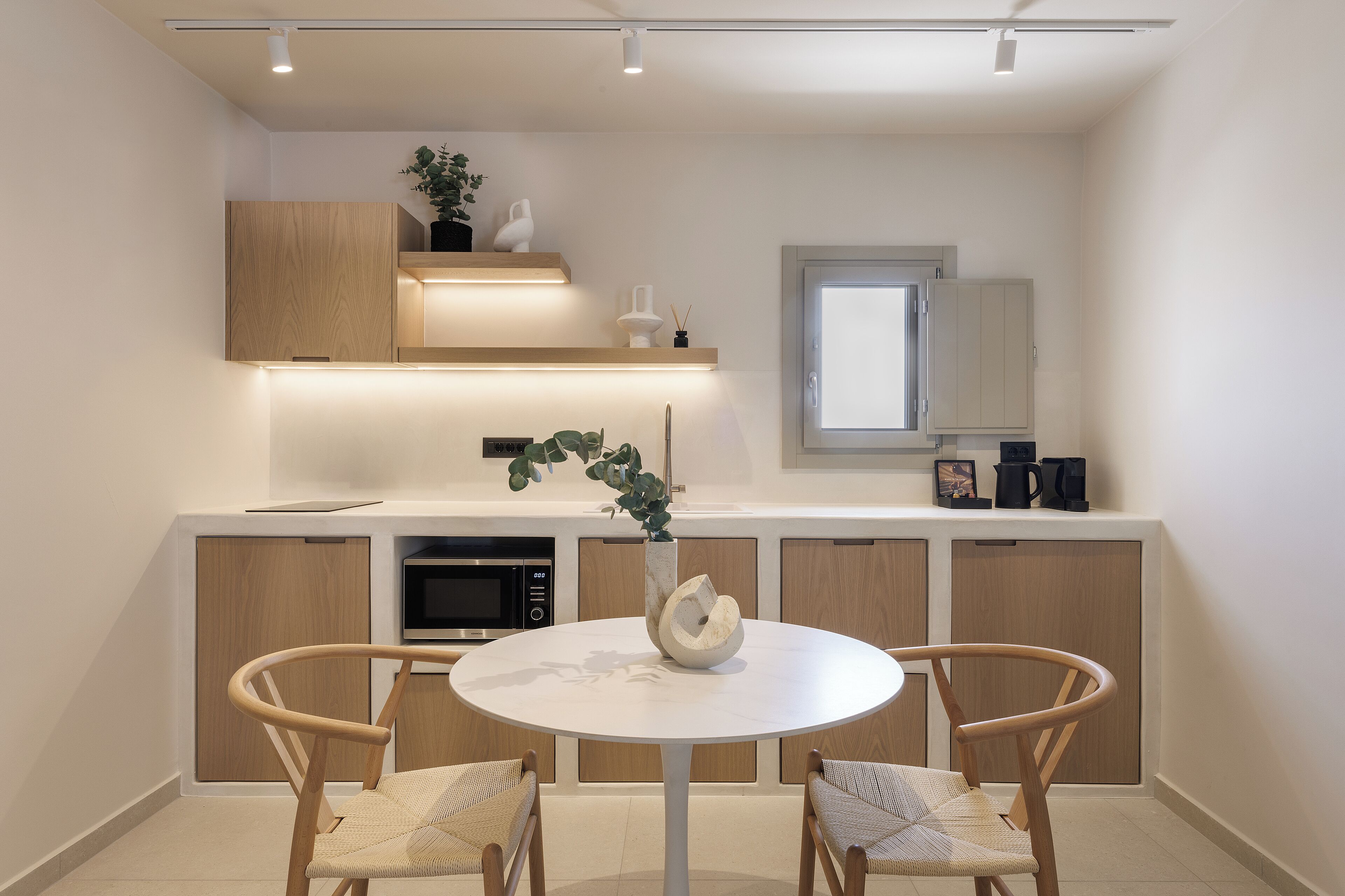 Une cuisine moderne avec des meubles en bois, un plan de travail blanc, une table de salle à manger ronde avec deux chaises et une plante en pot.