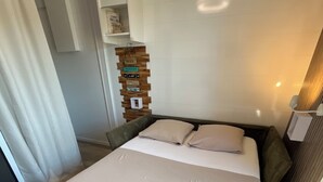 Un lit bien fait, avec une couette blanche et deux oreillers, une tête de lit en bois, une petite étagère en bois ornée d’objets décoratifs et un mur blanc agrémenté d’un luminaire.