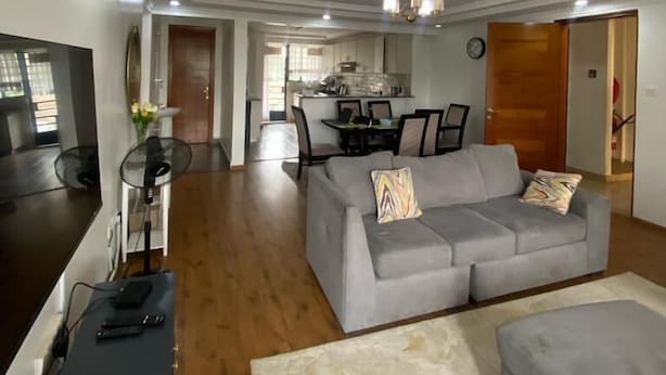A spacious living area with a grey sofa, a small ottoman, and a dining table with chairs. There is a kitchen in the background, a wooden floor, and a ceiling with recessed lighting. - Riverside Homes 6C (Nairobi)