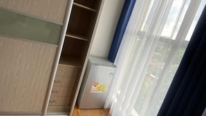 A room with a wooden floor, a wardrobe with multiple shelves, a small refrigerator, and a window with sheer curtains. - Riverside Homes 6C (Nairobi)