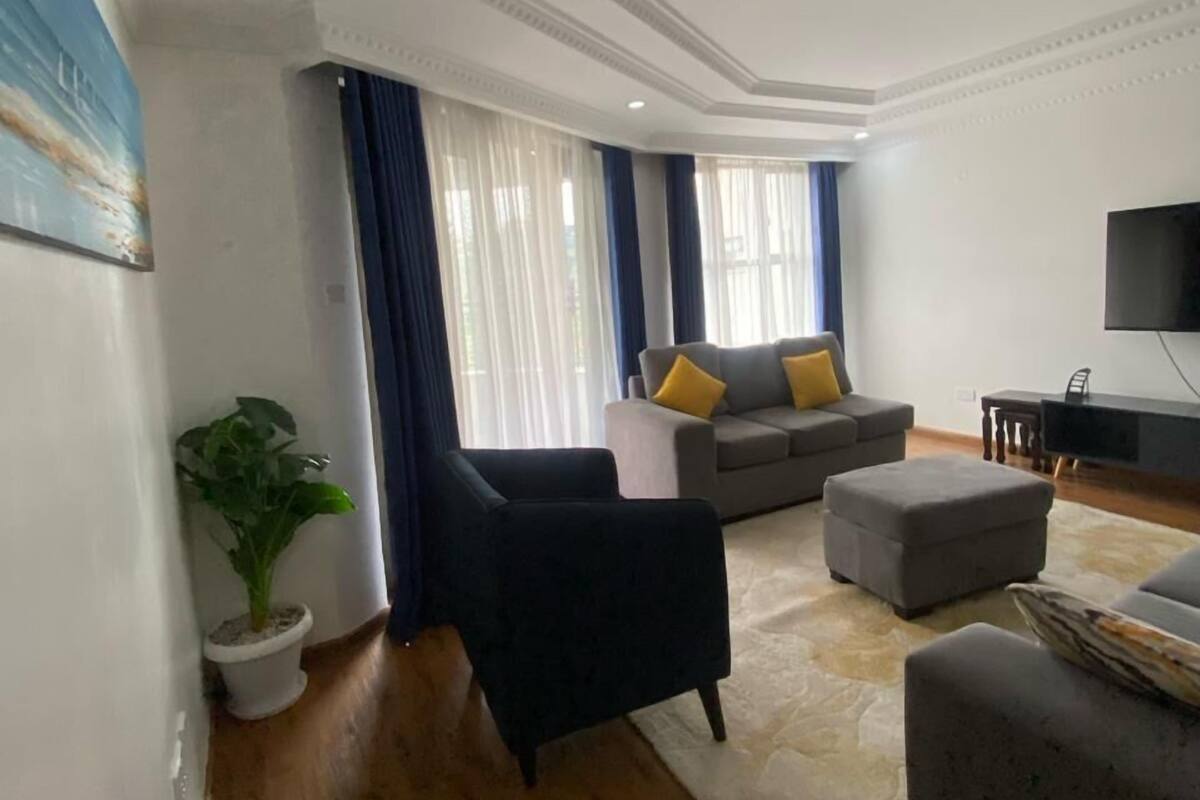 A modern living room with a grey sofa, a black armchair, and a grey ottoman. There is a flat-screen TV mounted on the wall, a potted plant, and a large window with curtains.