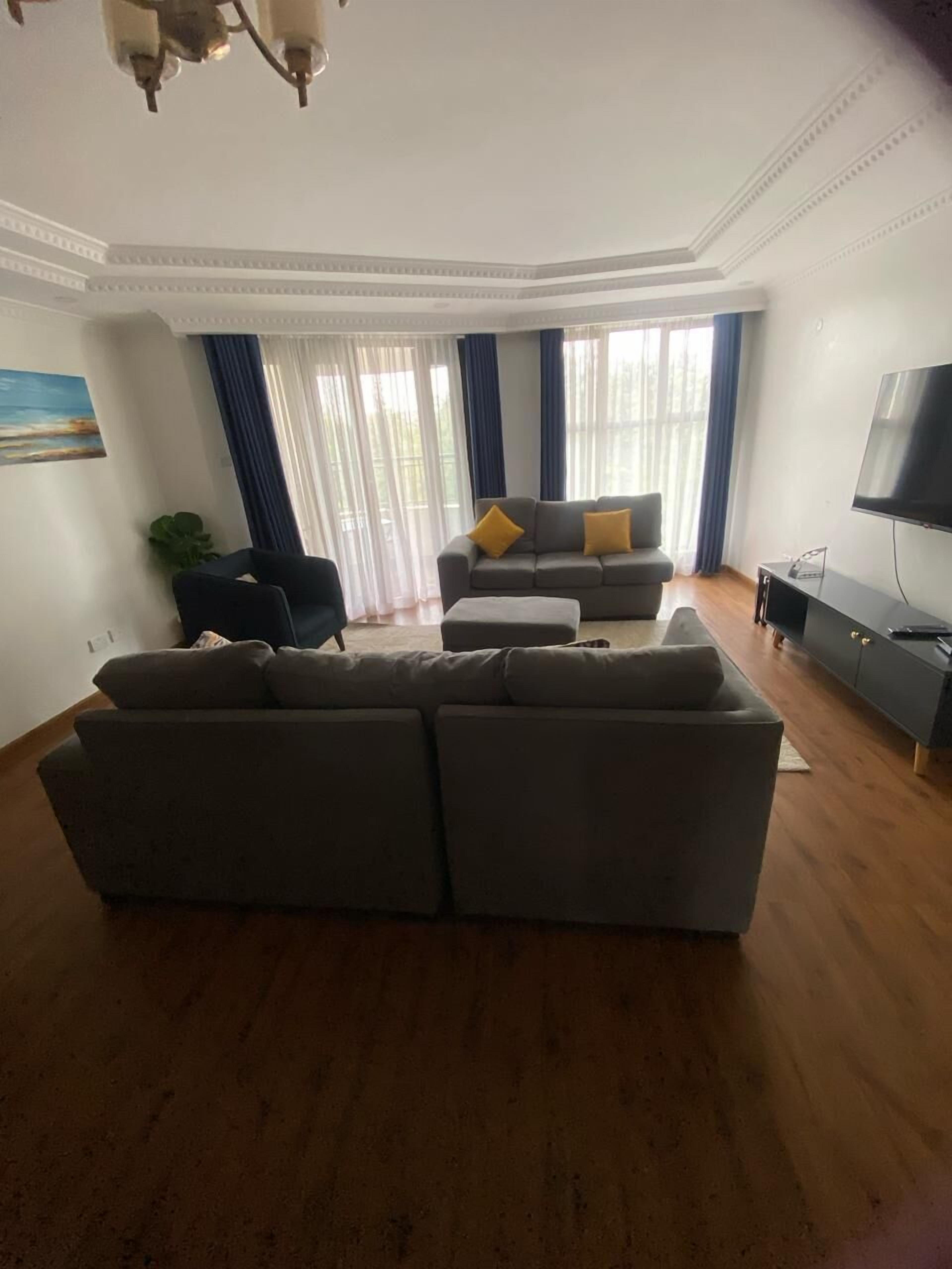 Een woonkamer met een grote grijze bank, een kleinere bank met gele kussens, een tv-meubel en een plafondlamp.