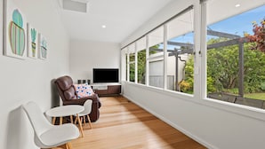 A modern living room with a large window, a leather sofa, a flat-screen TV, and a dining area with white chairs.