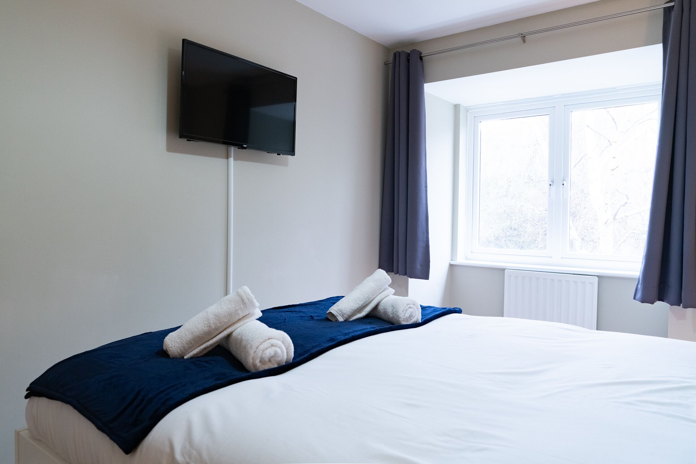 A hotel room with a bed, a flat-screen TV mounted on the wall, and a window with curtains.