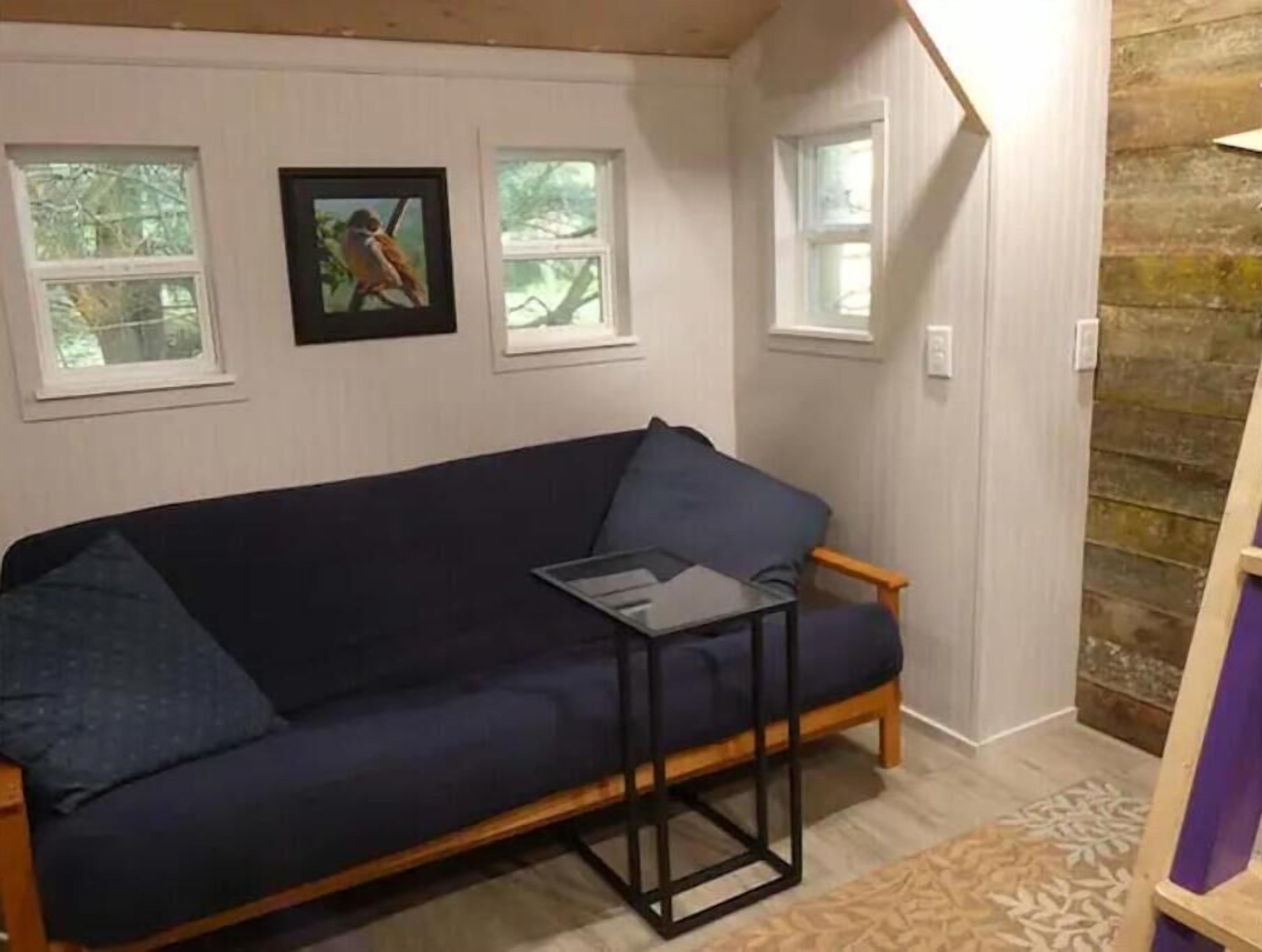 A room with a dark blue sofa, a small black table, and a patterned rug. There are three windows with white frames, and a picture is hanging on the wall.