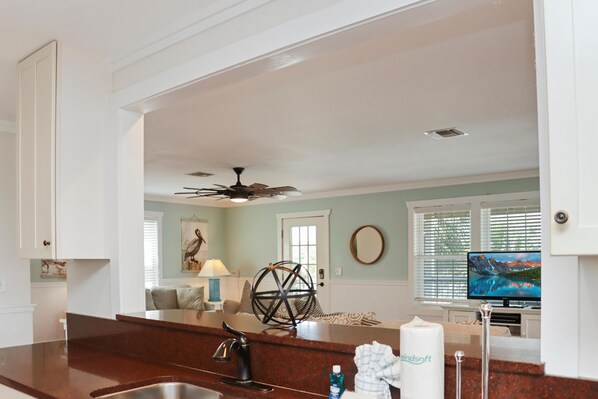 A kitchen with a dark brown countertop, a stainless steel sink, a ceiling fan, and a television in the background. - Cape Dreamin' (Port St. Joe)
