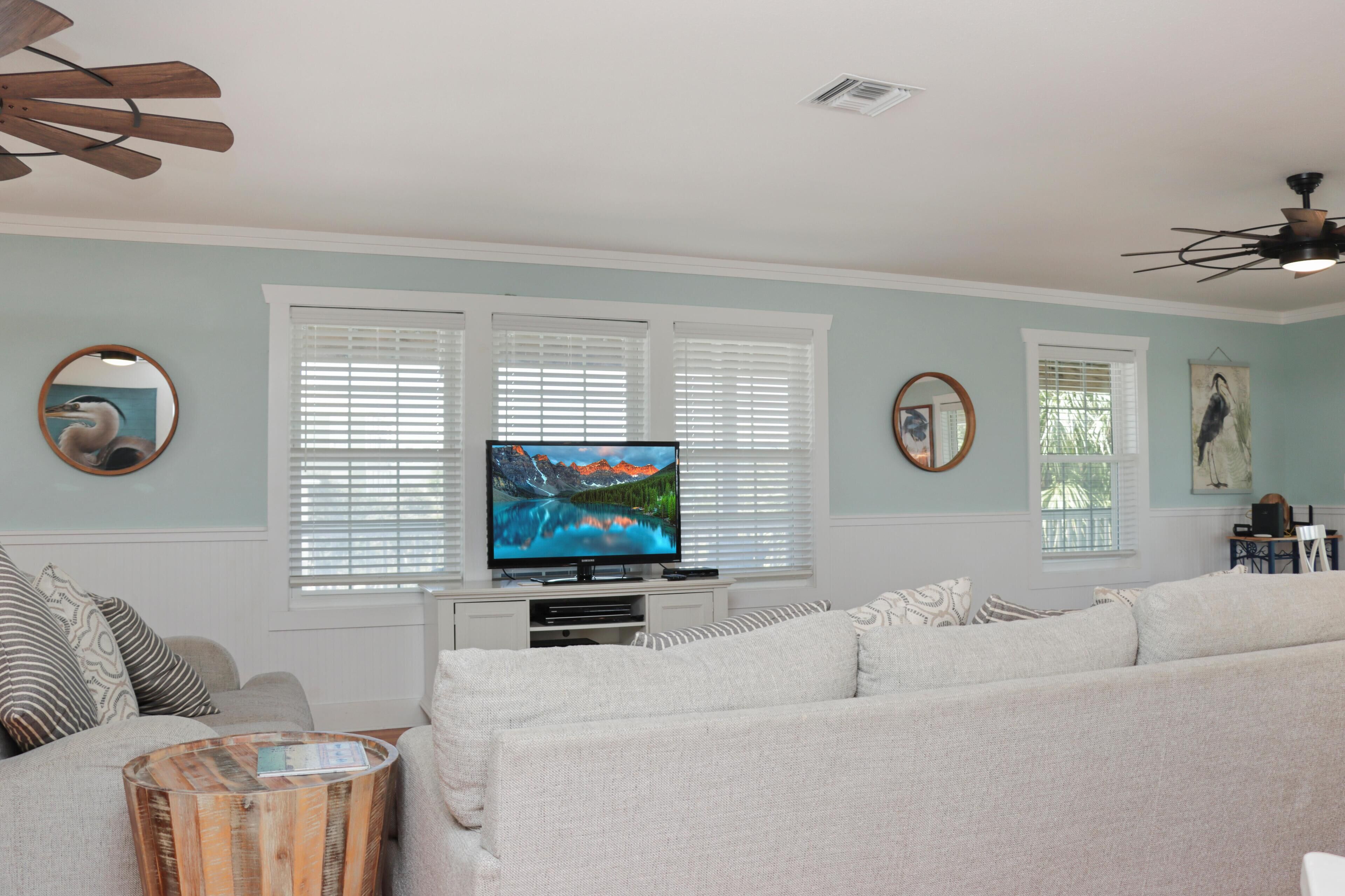 A living room with a large flat-screen TV, white sofas, a wooden coffee table, and a ceiling fan.