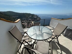 Ein Balkon mit rundem Tisch und zwei Stühlen, der Blick auf die Küste mit Hügeln und einem Gewässer gerichtet ist.