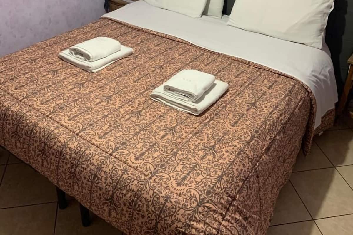 A neatly made bed with a patterned bedspread and two folded towels, a nightstand with a lamp, and a wall-mounted picture.