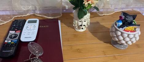 A wooden table with a red book, keys, two remote controls, a vase with flowers, and a container with snacks.