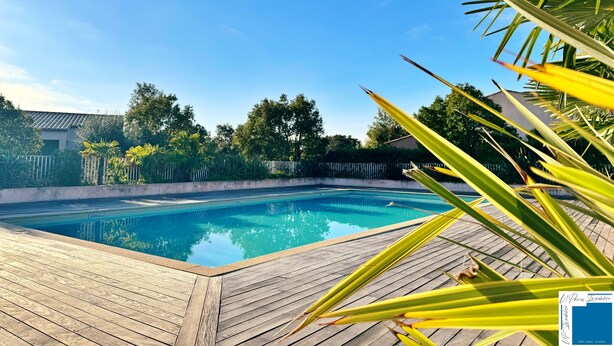 Une piscine dotée d’une terrasse en bois, entourée de verdure et surplombée d’un ciel bleu clair.