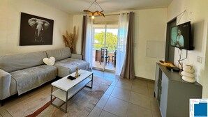 Un salon moderne avec un canapé gris, une table basse, un téléviseur fixé au mur et un balcon offrant une vue sur la verdure.