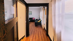 Un couloir au plancher de bois mène à un espace de vie comprenant un canapé et des coussins.