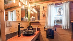 Baño de cabaña de madera con lavamanos, espejo y ventana con cortinas.