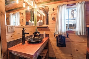 Une salle de bain rustique, aménagée dans une cabane en bois, avec un lavabo, un miroir et une fenêtre agrémentée de rideaux.