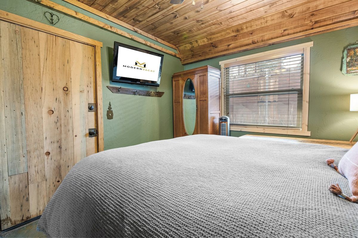 A bedroom with a wooden ceiling, a bed with a grey blanket, a wooden wardrobe, a television on the wall, and a window with blinds.