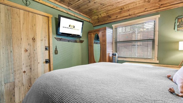 Un dormitorio con techo de madera, una cama con manta gris, un armario de madera, un televisor en la pared y una ventana con persianas.