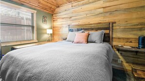 Un dormitorio con techo de madera, una cama con manta y almohadas grises, una mesita de noche con lámpara y una ventana con persianas.