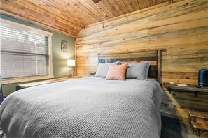 Une chambre avec un plafond en bois, un lit recouvert d’une couverture grise et agrémenté d’oreillers, une table de chevet avec une lampe, et une fenêtre avec des stores.