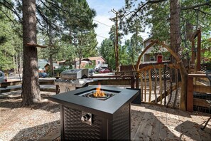 Un barbecue extérieur de forme carrée, avec une flamme visible, entouré d’arbres et d’une clôture en bois.