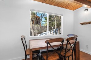 A dining area with a wooden table and chairs, a window with a view of trees and a building, and a white wall.