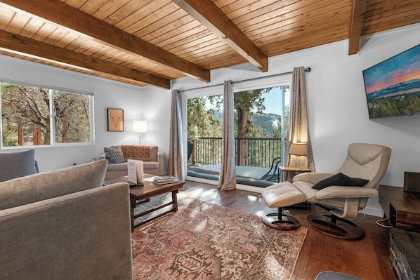 A modern living room with a wooden ceiling, large windows, a sofa, a chair, and a patterned rug.