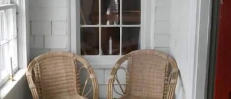 A room with two wicker chairs and a small round table, a large window, and a red door.