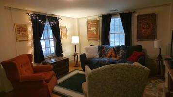 Una sala de estar acogedora con un sofá, un sillón y un pequeño tapete. Hay dos ventanas con cortinas, una lámpara sobre una mesita auxiliar y una pared con un cartel.
