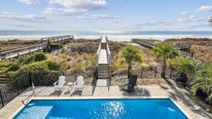 A pool area with a view of the beach, wooden deck, and lounge chairs.