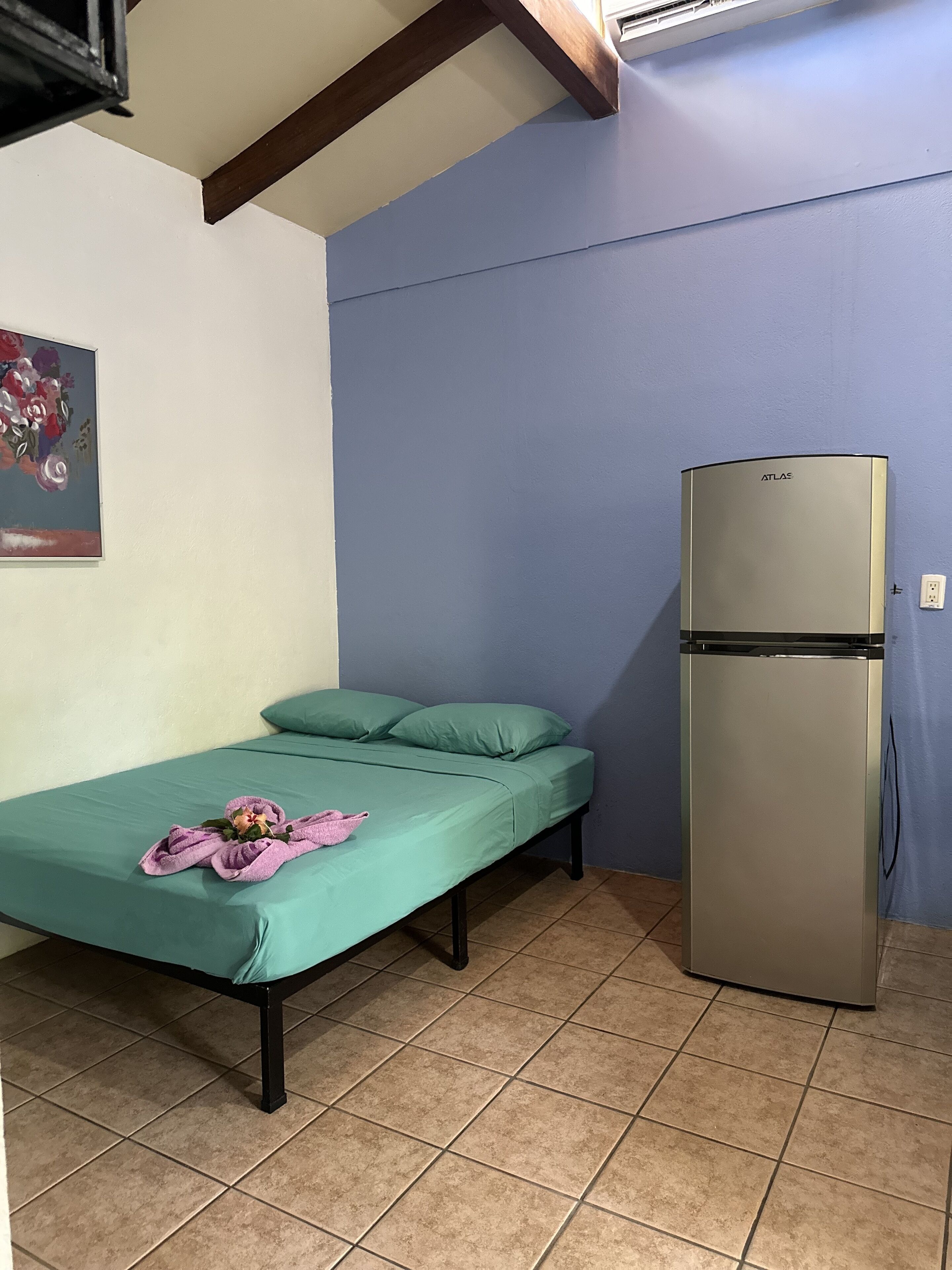 Una habitación pequeña con una cama, un colchón verde, una toalla rosa y un refrigerador.