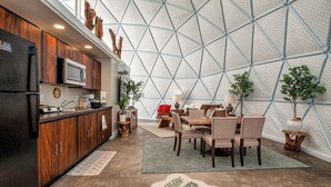 A modern kitchen with wooden cabinets and a microwave, a dining area with a table and chairs, and a large geometric wall structure. - Traverse City - Luxury on the Edge -13 (Williamsburg)