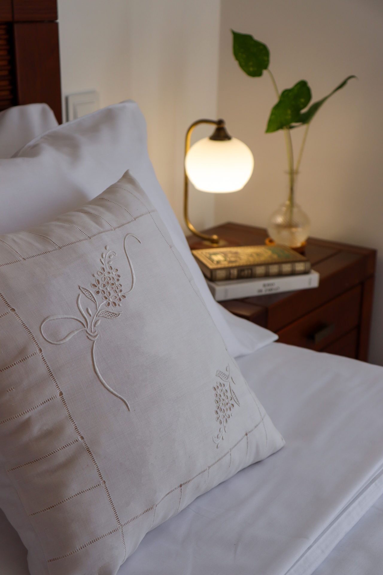Un lit bien fait, avec un oreiller orné de motifs floraux brodés, une lampe de chevet et une petite plante posée sur une table de chevet.