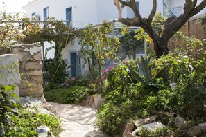 Un camí de pedra que condueix a un edifici blanc amb persianes blaves, envoltat de vegetació exuberanta i plantes florides.