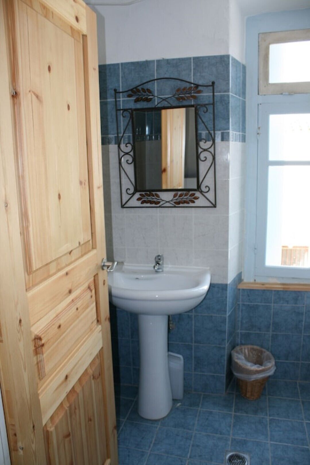 Een badkamer met een houten deur, een wastafel op een zuil, een spiegel aan de muur, blauwe tegels en een raam.