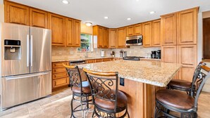A kitchen with wooden cabinets, stainless steel appliances, a granite island with bar stools, and recessed ceiling lights.