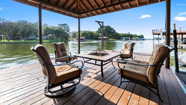 A covered outdoor seating area with a table and chairs, overlooking a body of water with boats and buildings in the background.