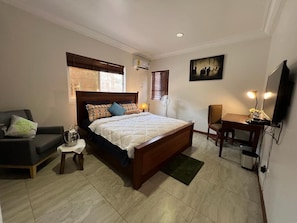 A bedroom with a bed, a chair, a desk, a TV, and a window with blinds.