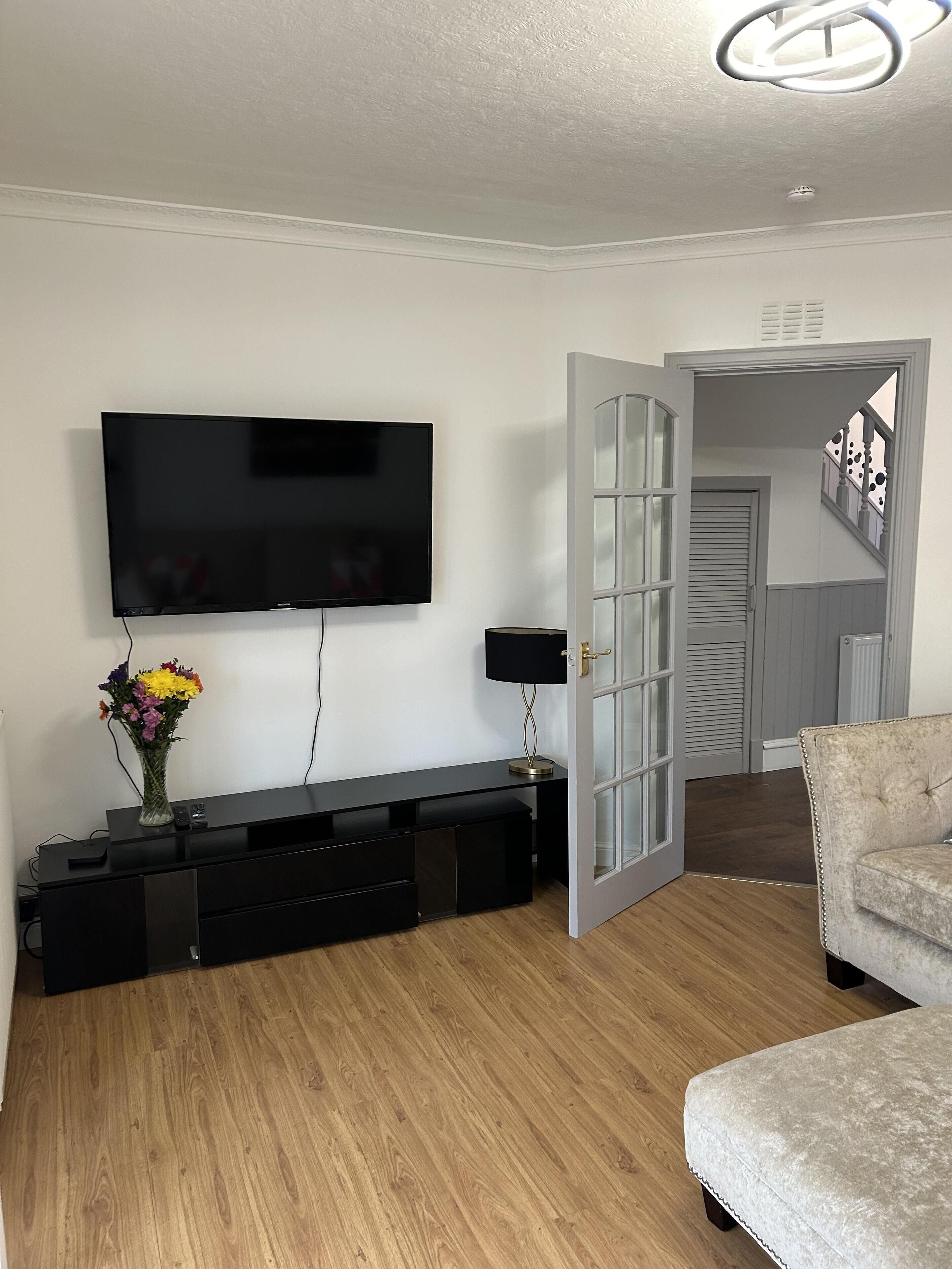A modern living room with a flat-screen TV, a black entertainment unit, a vase with flowers, and a grey cushioned bench.