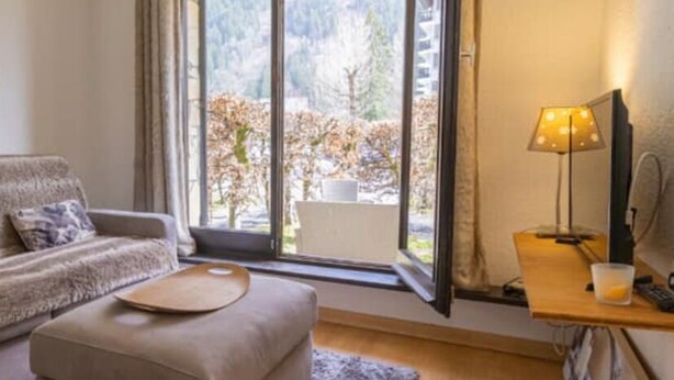 Un accogliente soggiorno con un divano, un poggiapiedi rotondo, un tavolino in legno con una lampada e vista sugli alberi attraverso un'ampia finestra.