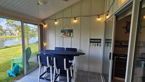 Un espacio al aire libre cubierto con una mesa y sillas, un vidrio que da a una cafetería, y una vista de un lago y edificios al fondo.