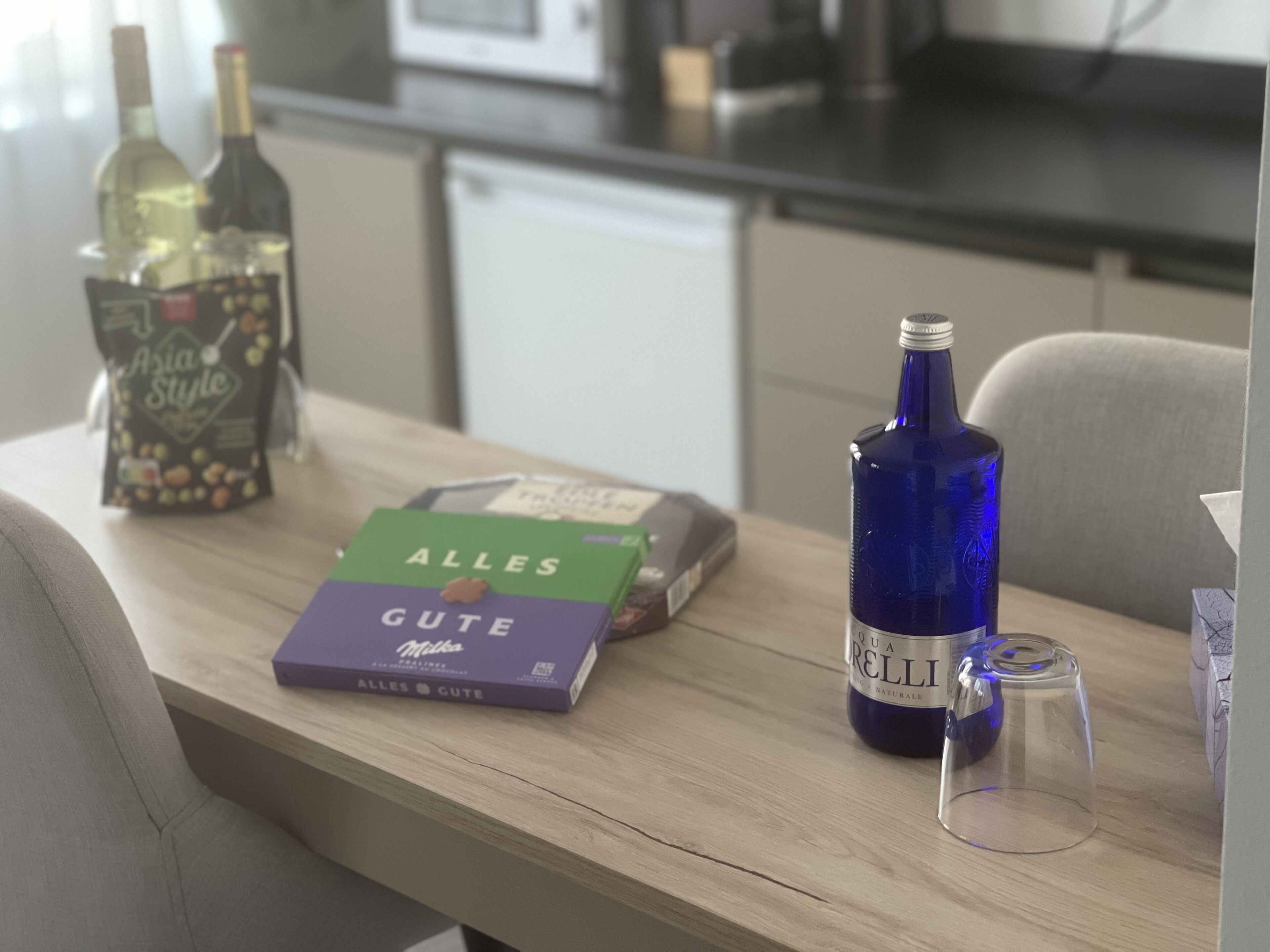 Una mesa de madera con una caja de bombones 'Alles Gute', una botella de vino 'Qua Relli' y una copa de vino.