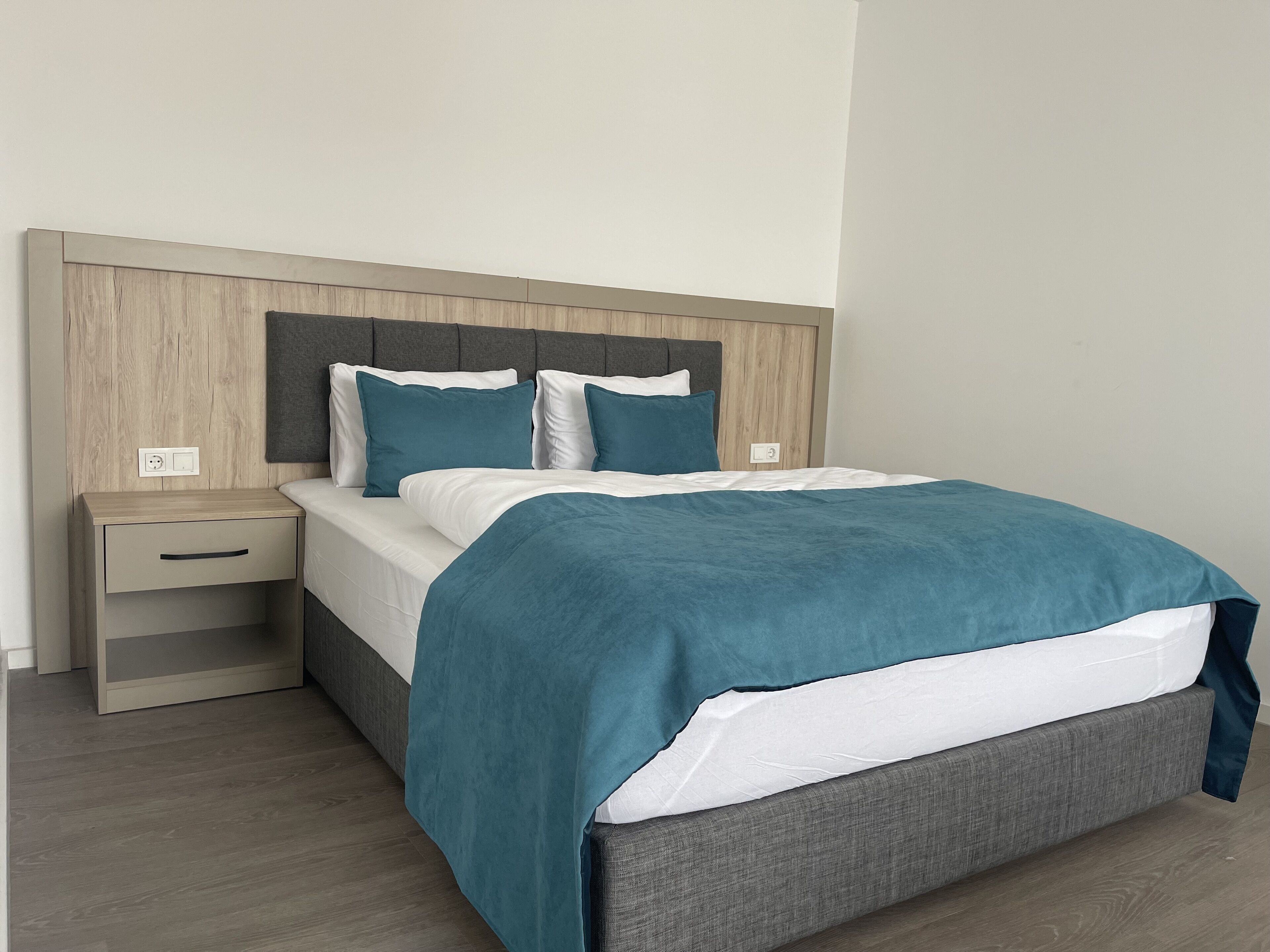 A modern bedroom with a wooden headboard, a single bed with a teal blanket, and a nightstand with a drawer.
