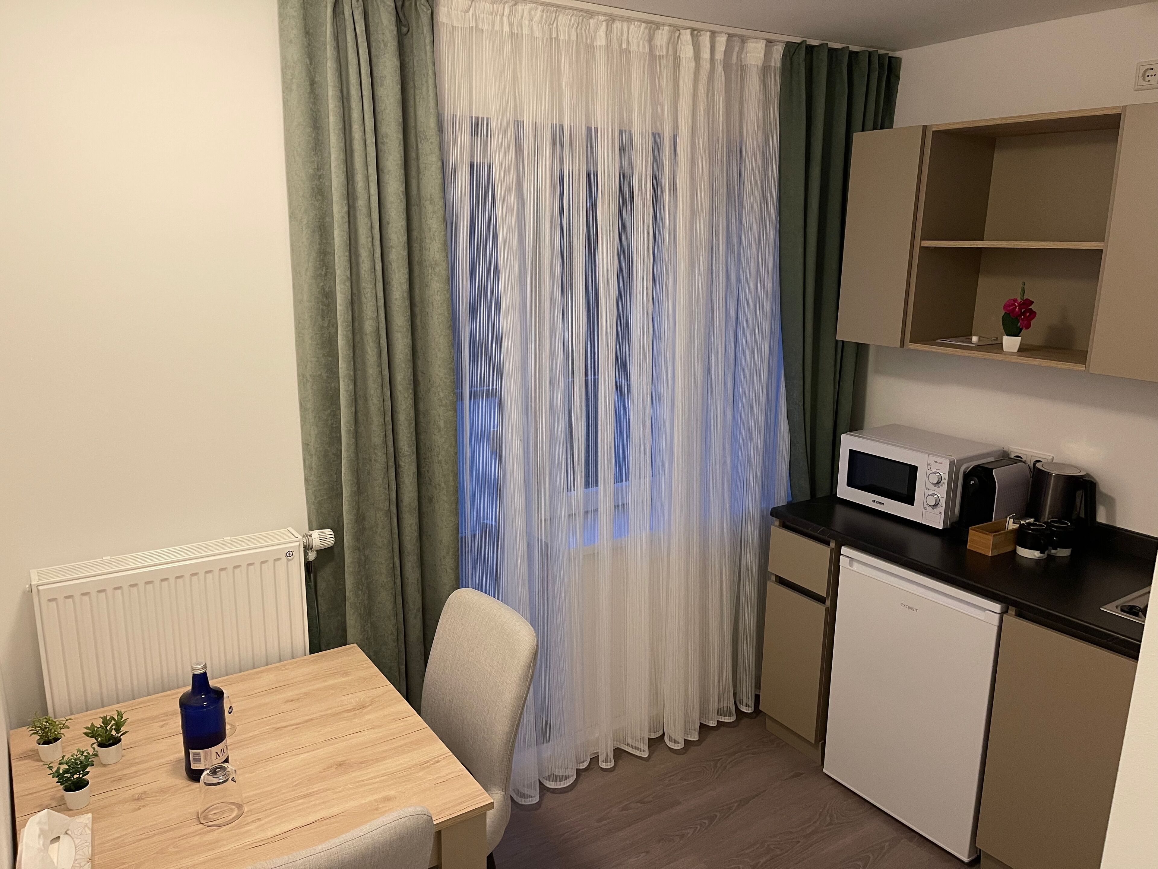 A small kitchenette with a microwave, coffee maker, and a mini fridge. There is a table with a bottle and glass, and a chair. The room has a window with sheer curtains.