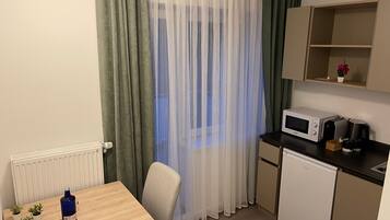A small kitchenette with a microwave, coffee maker, and a mini fridge. There is a table with a bottle and glass, and a chair. The room has a window with sheer curtains.