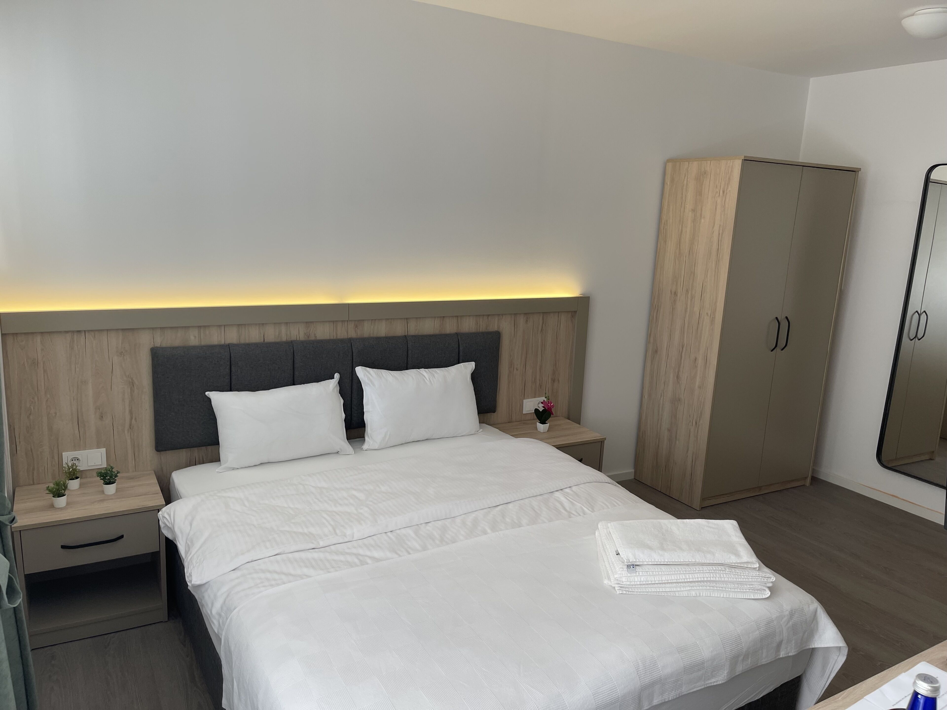 A hotel room with a large bed, a wooden wardrobe, a nightstand with a lamp, and a mirror.