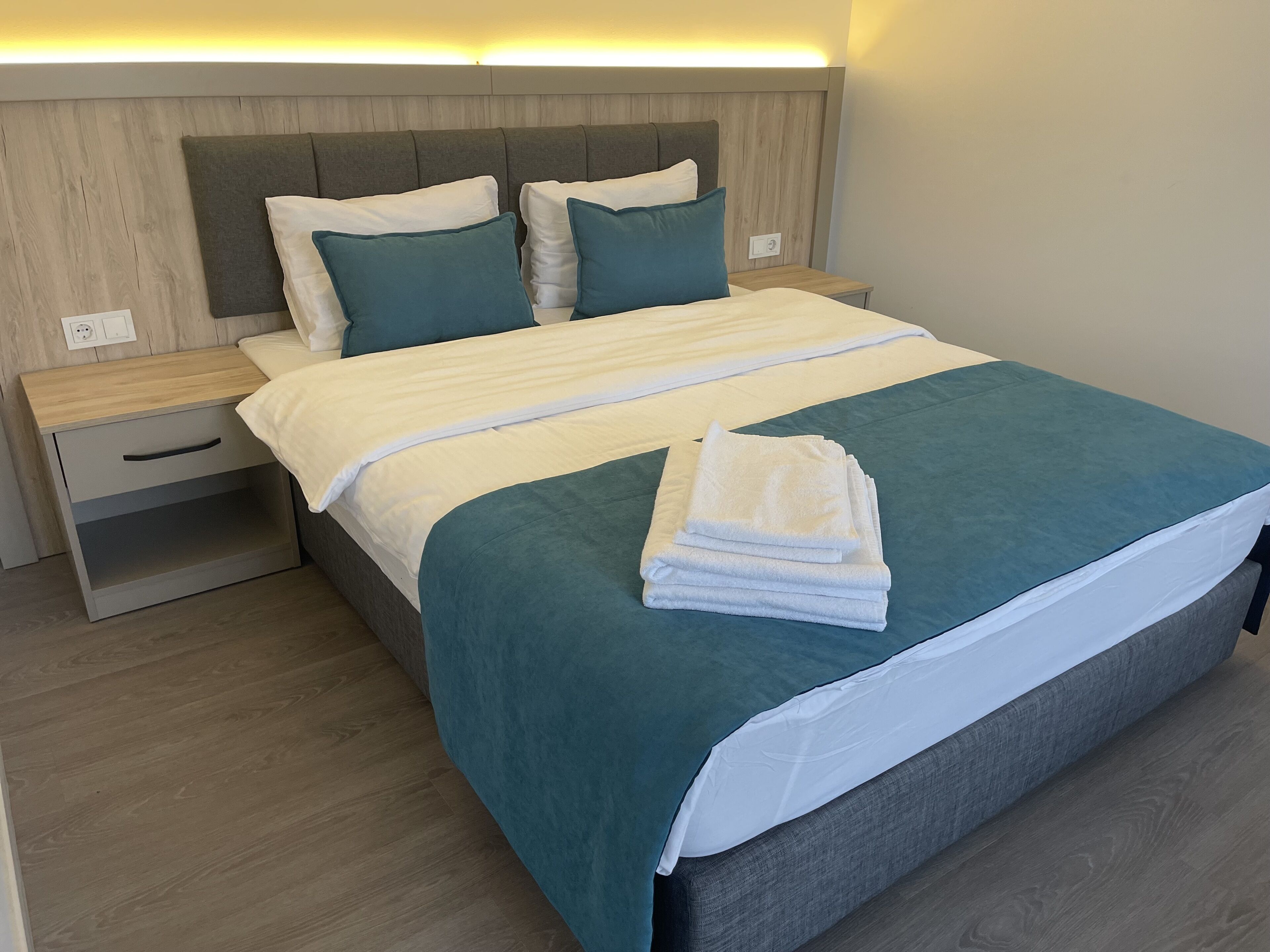 A neatly made bed with a teal bedspread and white linens, two pillows, and a folded white towel on the bed.
