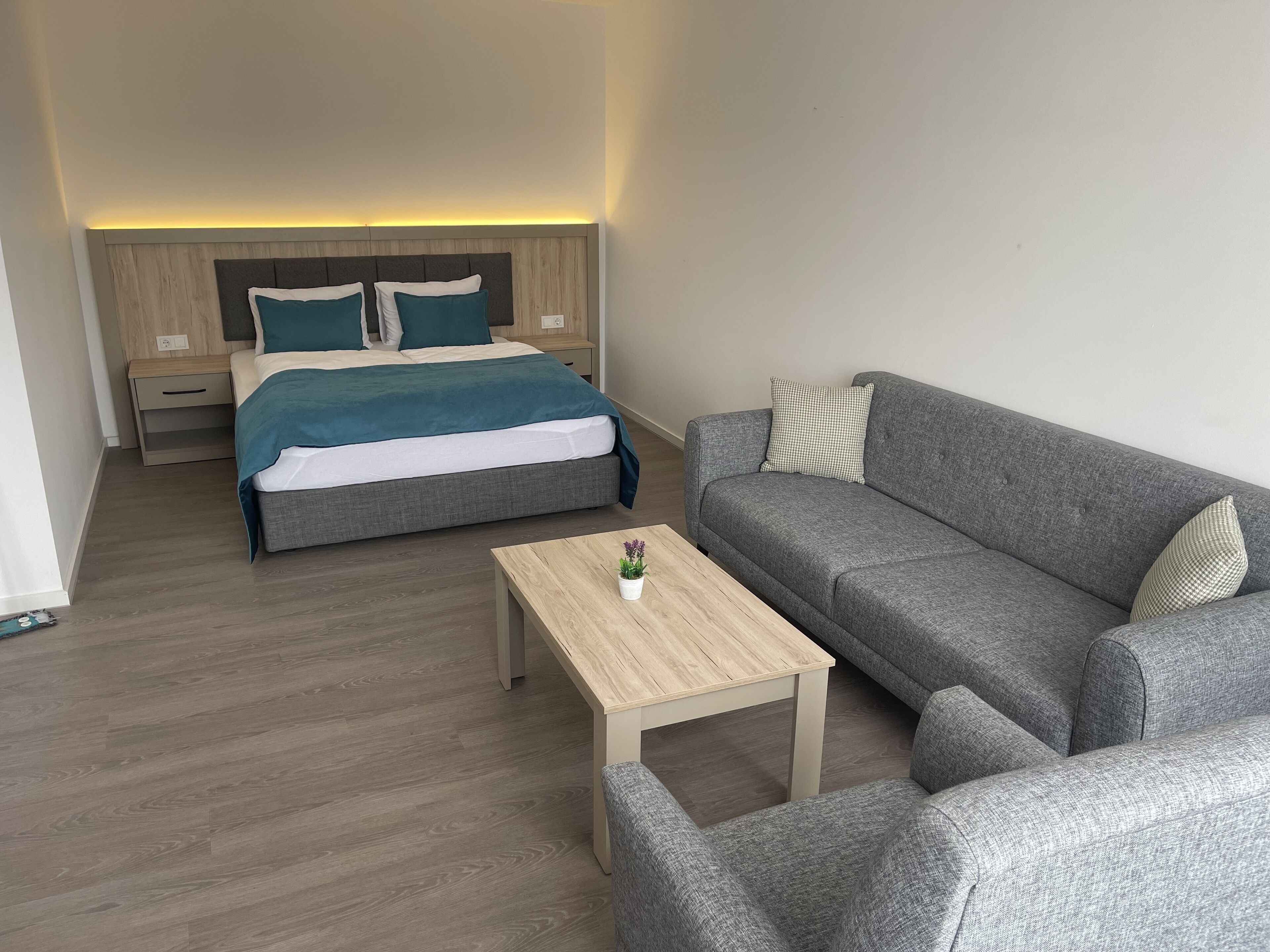 A modern hotel room with a bed, a grey sofa, a wooden coffee table, and a nightstand.